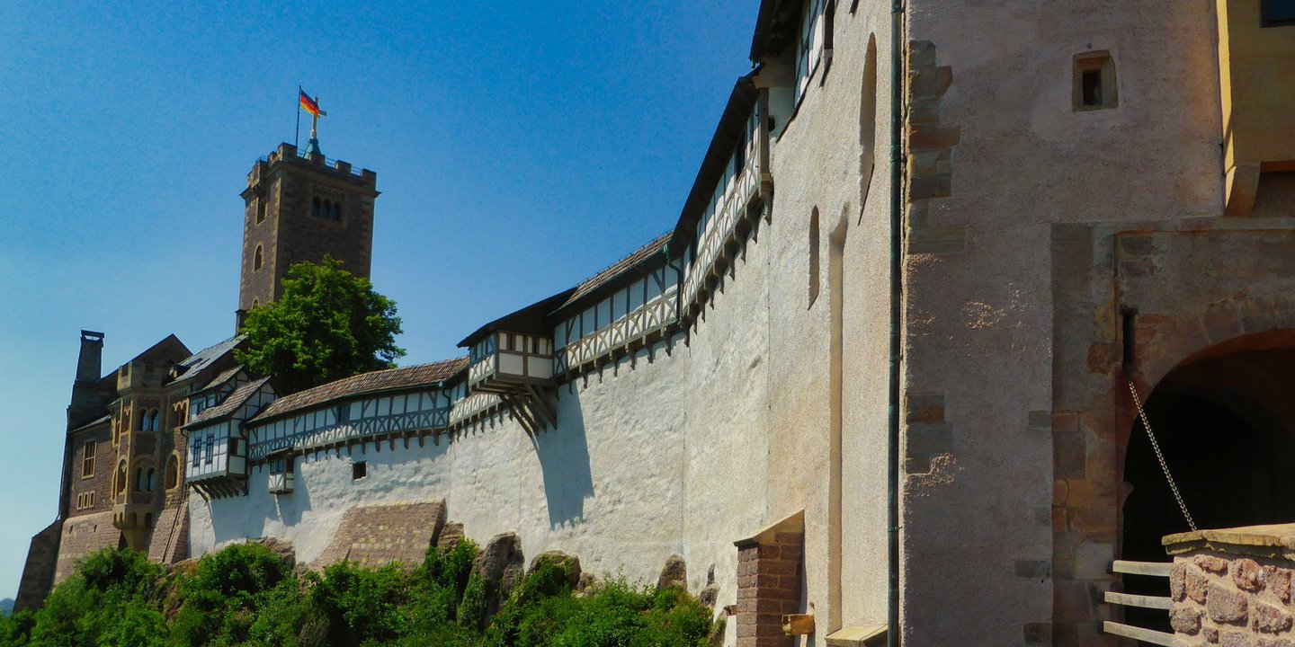 Bild entlang einer Burgmauer im Sonnenschein. Am Ende der Mauer steht ein Turm mit Deutschlandflagge.